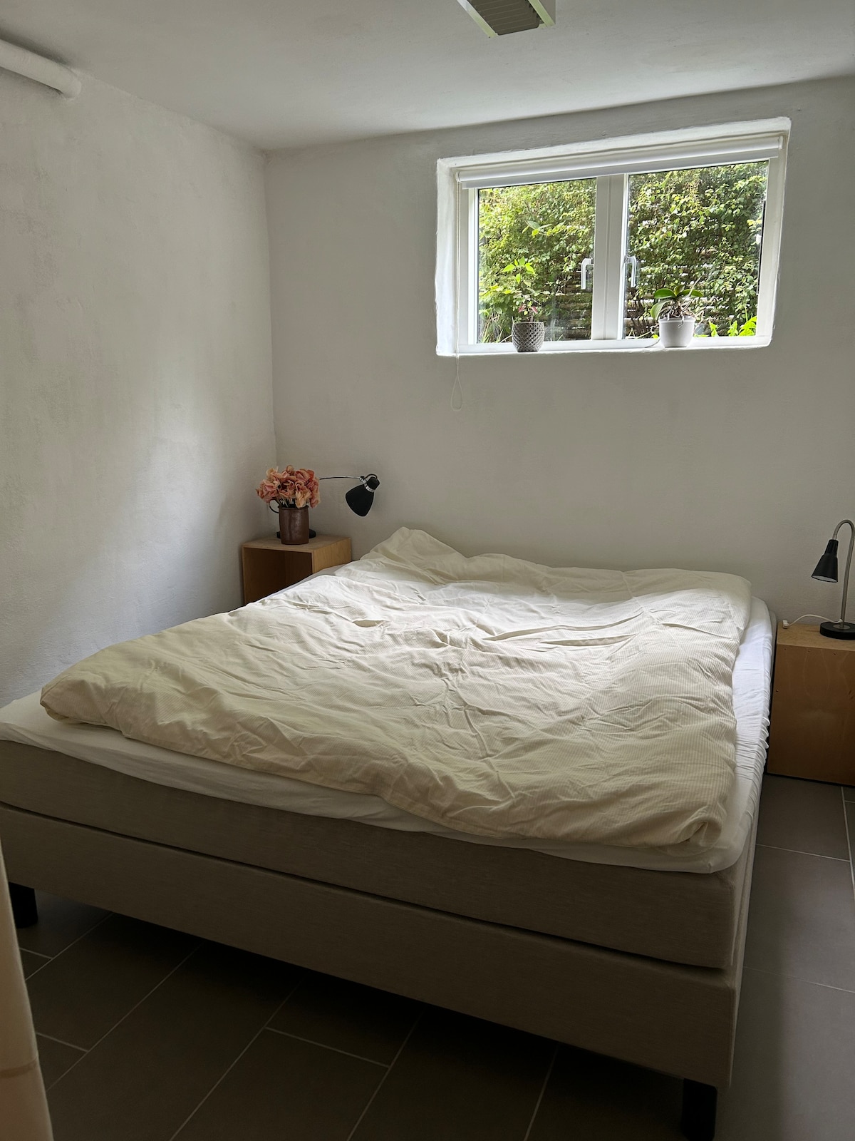 A bright bedroom contains a spacious bed with a light-colored duvet. A window is positioned above the bed, bringing in natural light and revealing a view of greenery outside. A bedside table with a small lamp and a floral arrangement adds a calming touch.