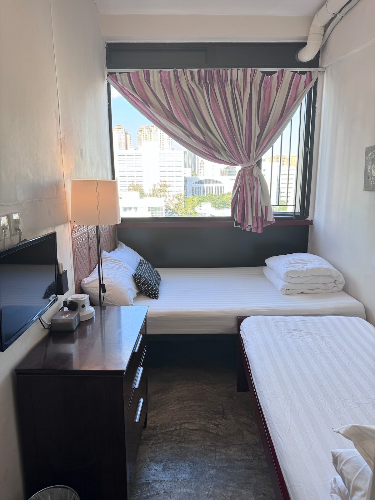 A room with two beds is seen, positioned closely beside each other. A window offers a view of the city skyline, partially covered by striped curtains. A bedside table with a lamp and a small television is present, enhancing the functionality of the space.