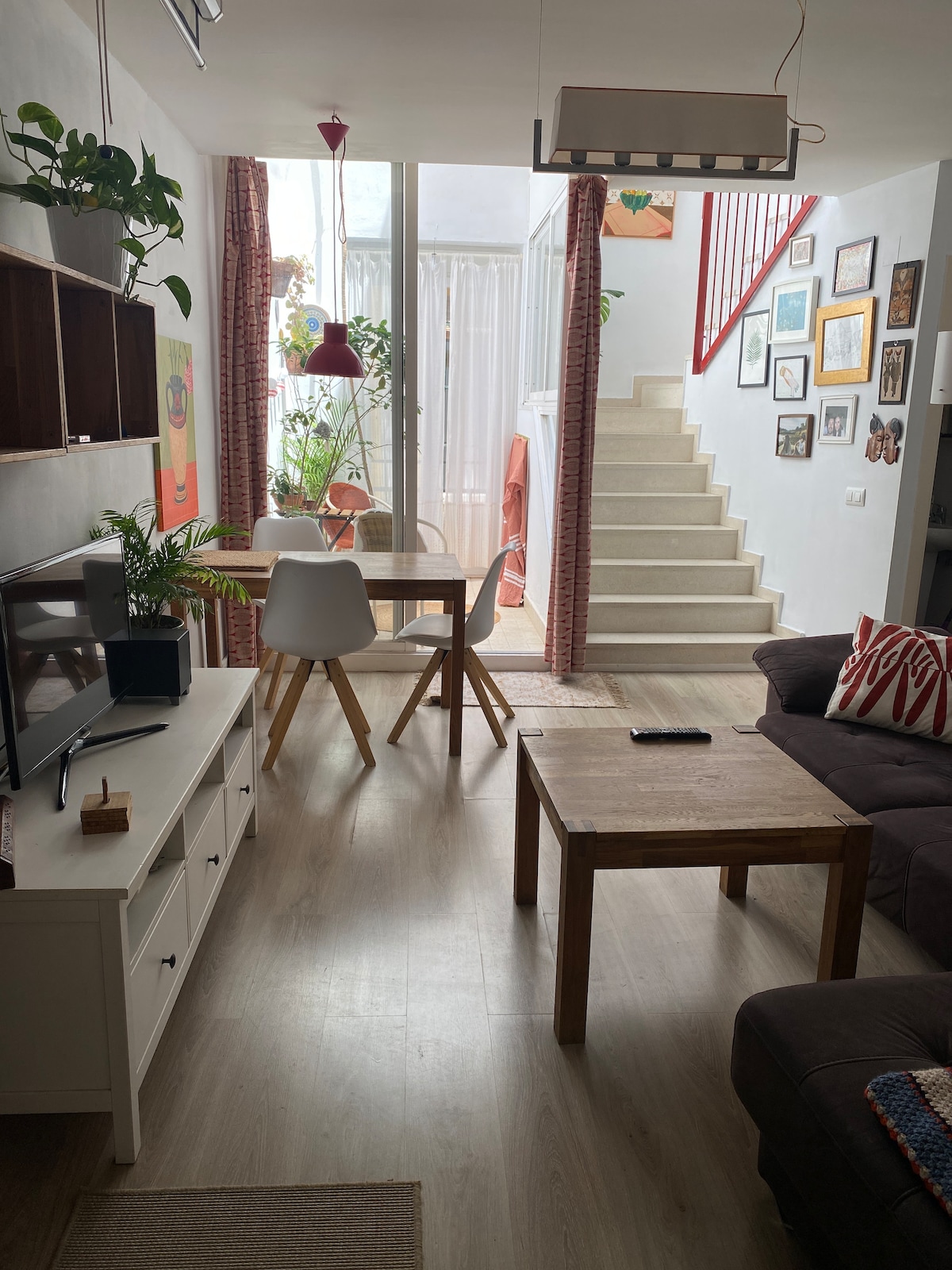 A bright living area features a dark sofa and wooden coffee tables. A dining space with light chairs is visible alongside greenery. Light filters through sheer curtains, illuminating a staircase leading to the upper level adorned with various framed artworks.