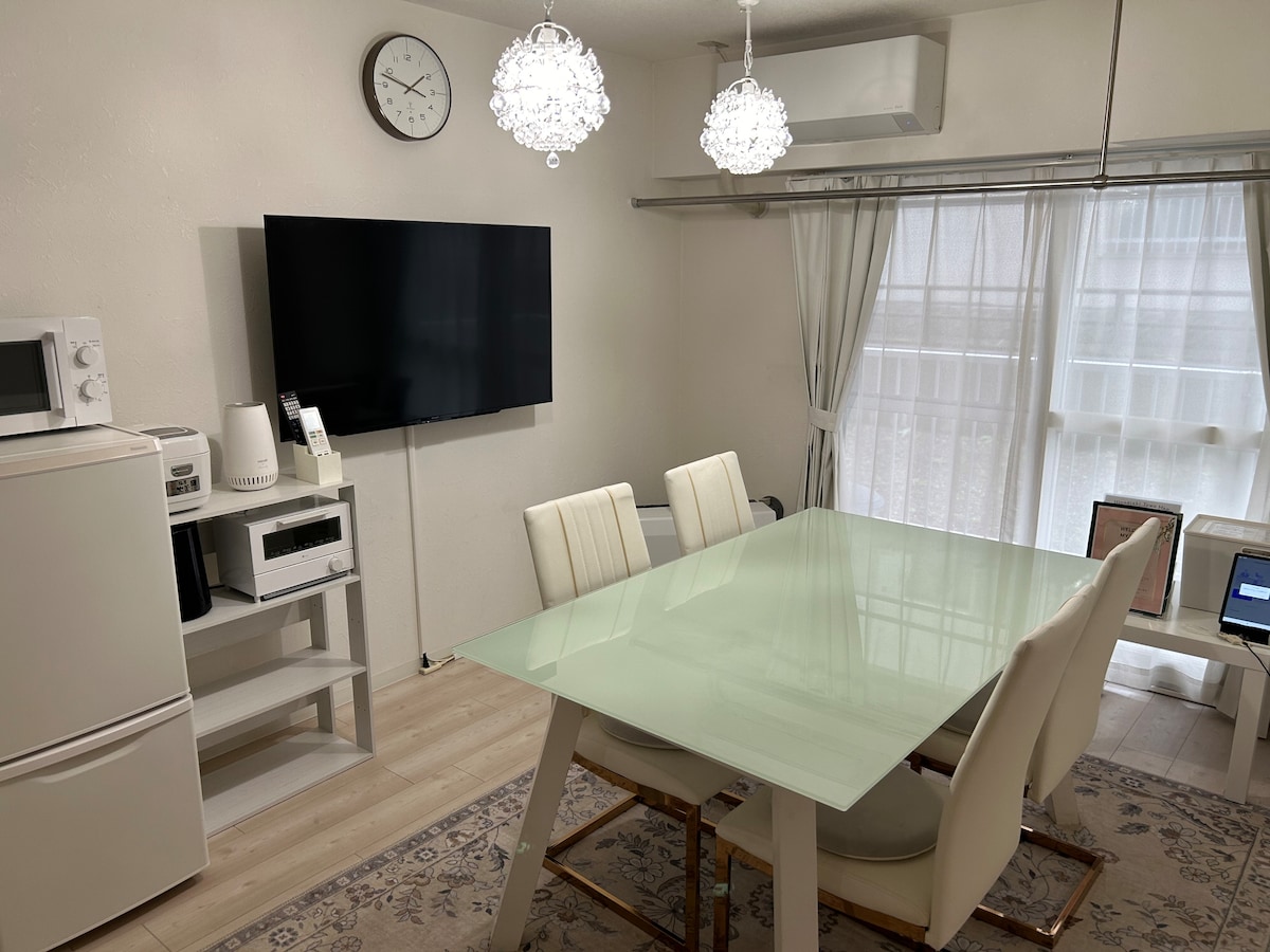 A spacious dining area features a large glass-top table surrounded by four white chairs. A modern television is mounted on the wall, with various kitchen appliances, including a microwave and a refrigerator, located nearby. Natural light filters through the sheer curtains.