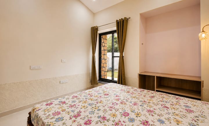 Une chambre lumineuse et confortable avec un grand lit, de la literie douce à fleurs, un éclairage chaleureux et une grande fenêtre donnant sur la verdure, créant une atmosphère paisible et relaxante.