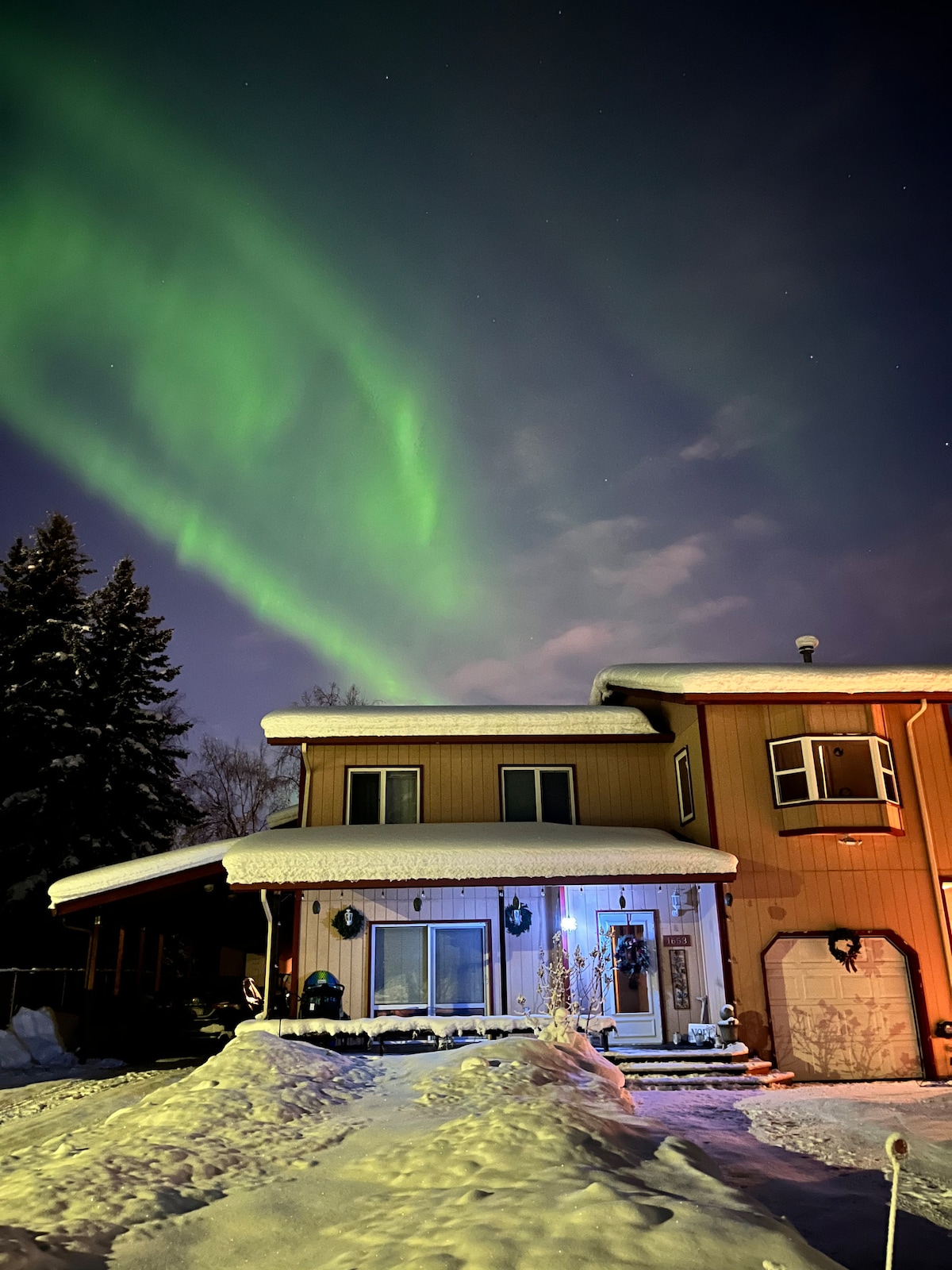 A two-story building is illuminated under a night sky, with vibrant green auroras dancing overhead. Snow blankets the ground and the roof, while subtle outdoor lighting highlights the entrance, adorned with seasonal decorations. Tree silhouettes frame the scene, enhancing the tranquil atmosphere.