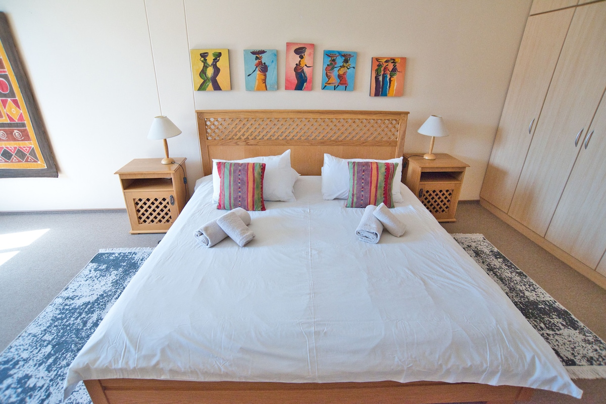 A spacious sleeping area is presented with a large bed topped with crisp white linens. Decorative pillows add color to the bedding. Two bedside tables with lamps flank the bed, while vibrant artwork depicting figures adorns the wall above.