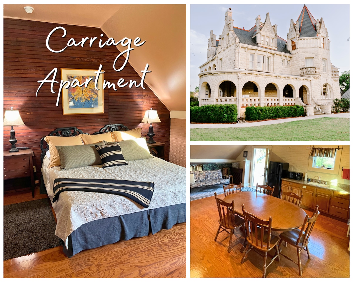 The image displays a cozy bedroom featuring a spacious bed adorned with decorative pillows and two bedside lamps. In the background, the historic carriage house is showcased, highlighting its unique architecture. On the right, a dining area is visible, featuring a large table surrounded by wooden chairs and a fully equipped kitchen.