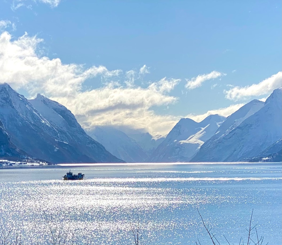 A serene view of snow-capped mountains flanks a shimmering fjord, with sunlight reflecting on the water's surface. A boat can be seen gliding gently across the expanse, contributing to the tranquil scene within the expansive landscape.