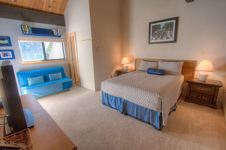 Recámara tipo loft con cama, futón, ventana, mesas de cabecera, lámparas, TV en el borde y techo alto.