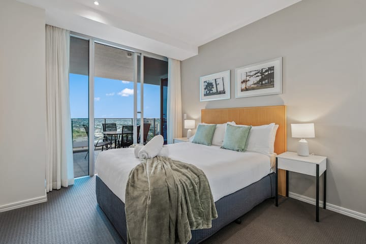 Rest easy in this inviting master bedroom featuring a plush bed adorned with crisp white linens and accent pillows, bedside tables with reading lamps, and direct sliding door access to a private balcony with expansive outdoor views.