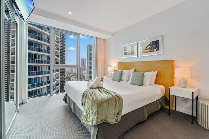 Wake up to stunning urban views in this bright master bedroom, featuring a plush king-sized bed, stylish soft green throw pillows, nightstands with lamps, and floor-to-ceiling windows overlooking the high-rise cityscape.