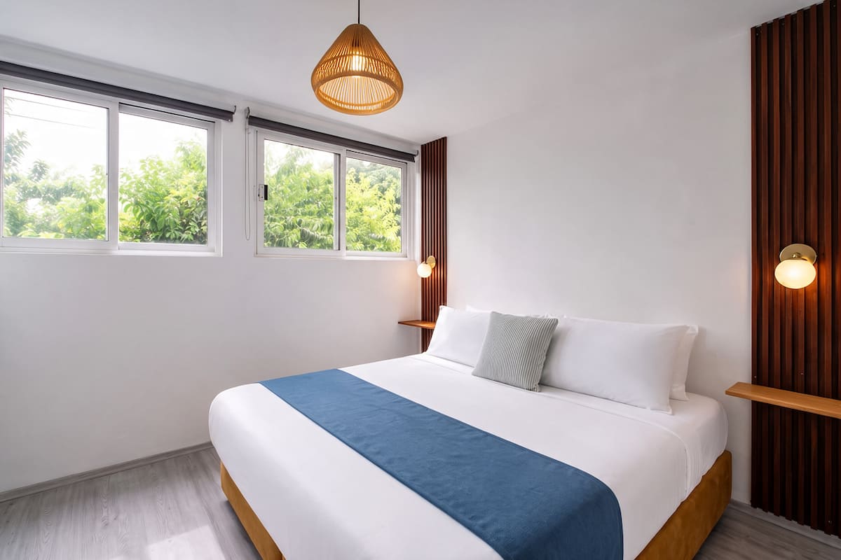 Recámara en Casa Mérida con muro de madera en listones, lámpara de ratán y ventanas hacia los árboles