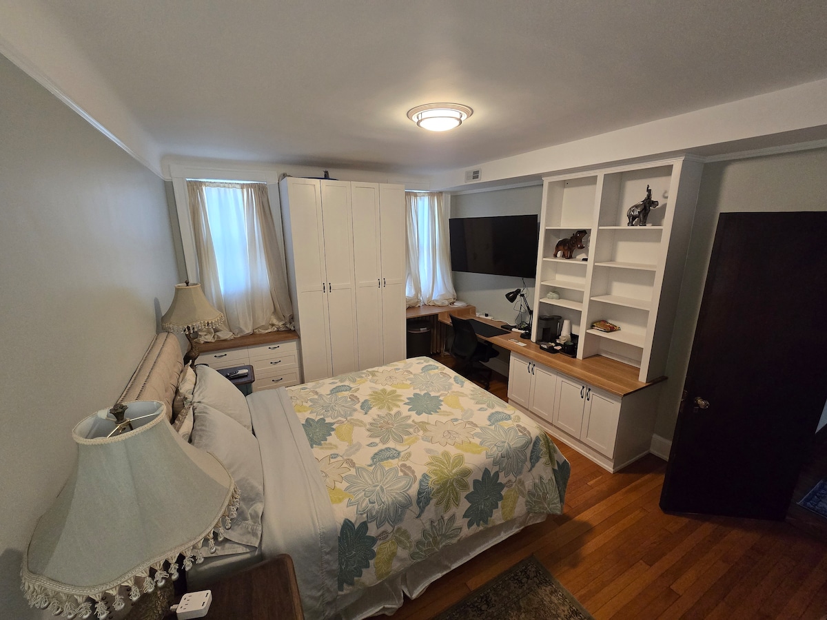 A private room features a queen-sized bed adorned with floral bedding. A spacious desk and chair are positioned against the wall near a mounted TV. Two large windows with light-draping curtains provide ample natural light. Built-in closets and shelves offer additional storage.