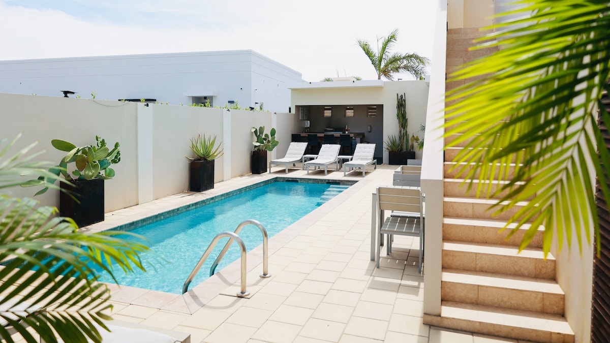 A serene pool area is bordered by tall, green plants and lined with lounge chairs. An outdoor dining setting can be seen adjacent to the pool, while steps lead to upper areas, creating a spacious and relaxing environment.
