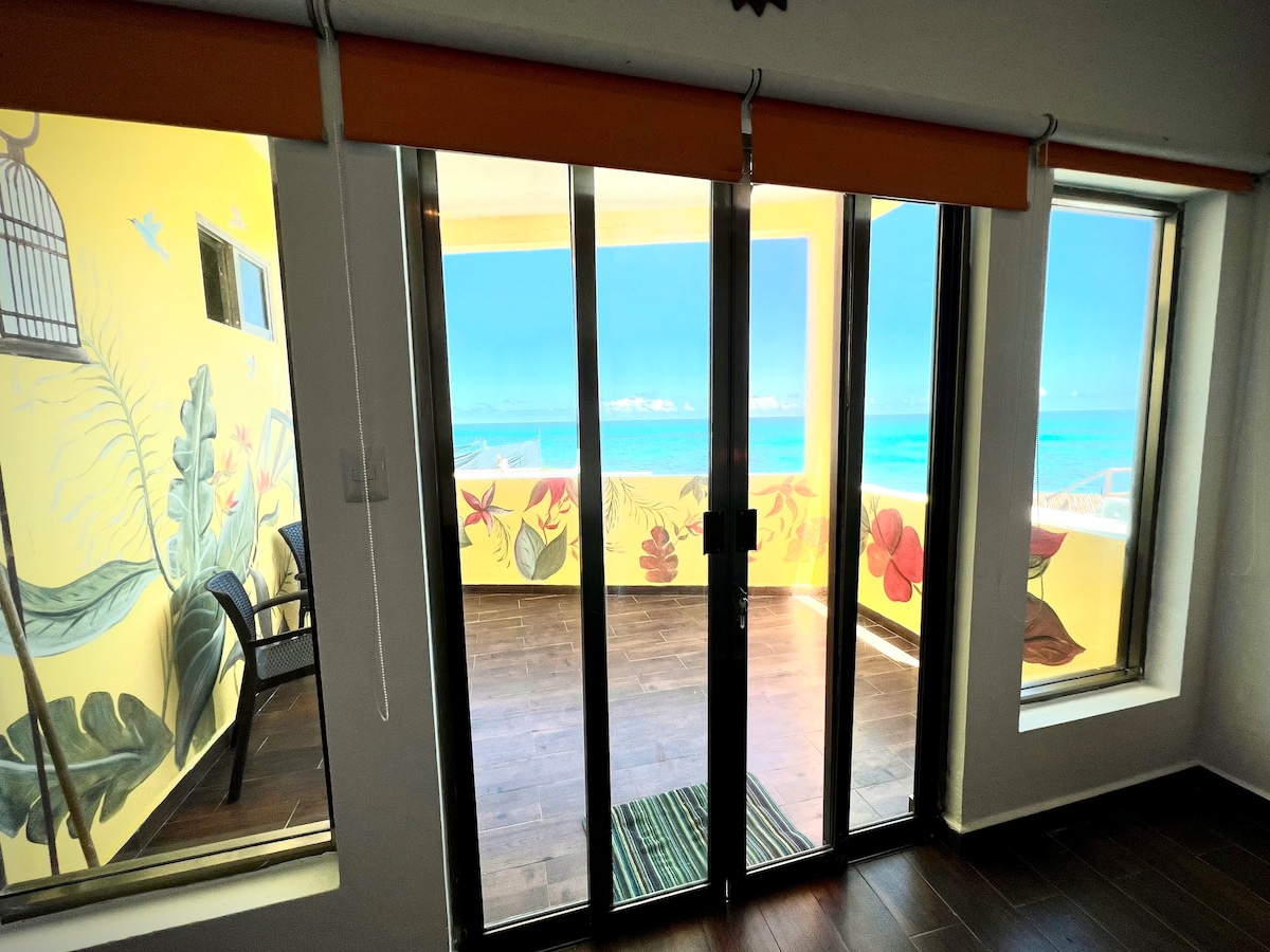 Sliding glass doors lead to a private balcony with a vibrant mural, offering views of the ocean. The space is enhanced by a warm, natural light filtering in, with a cozy seating area visible beside the doors on a polished wooden floor.