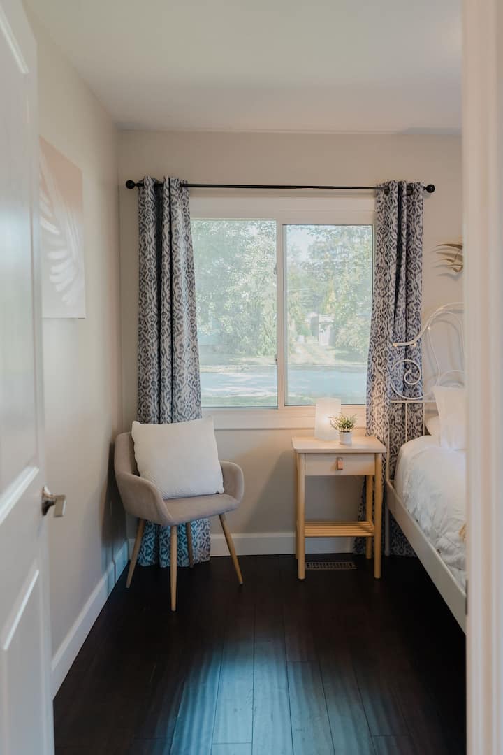 Chambre lumineuse d'inspiration côtière avec un lit en fer forgé blanc, un fauteuil confortable, des rideaux occultants à motifs, un éclairage de chevet chaleureux, des planchers en bois foncé et une grande fenêtre offrant de la lumière naturelle.