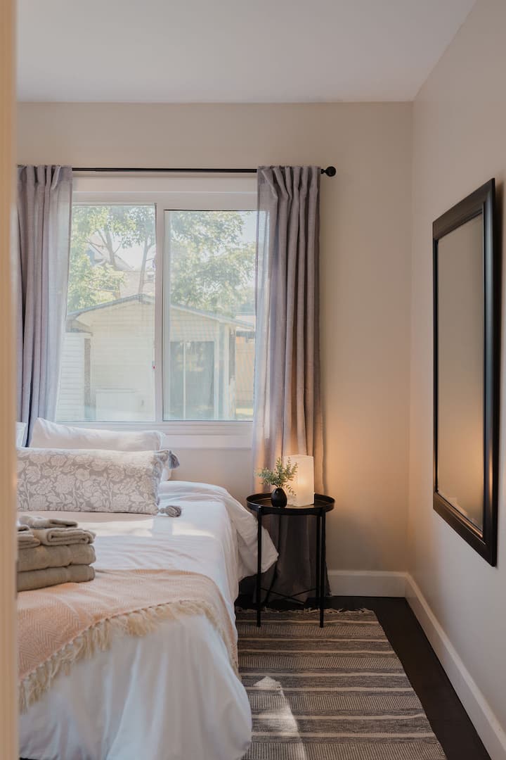 Chambre lumineuse et confortable avec de la literie blanche impeccable sur un lit King, des touches de rose pâle, des coussins décoratifs, des rideaux filtrant la lumière, deux tables de chevet avec un éclairage chaleureux, un miroir pleine longueur et de la lumière naturelle.