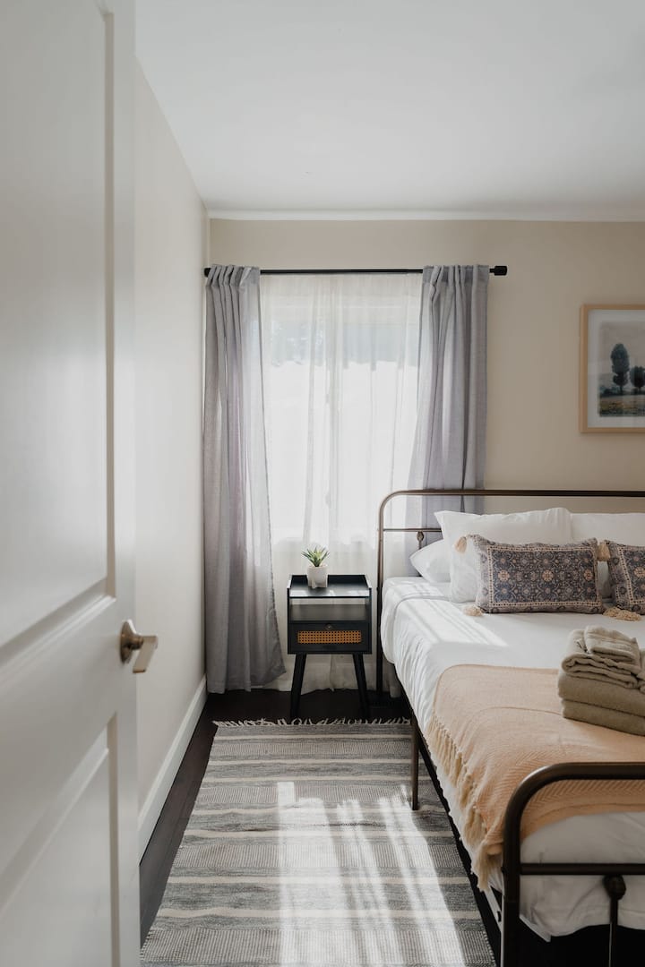 Chambre lumineuse et confortable avec un cadre de lit grand lit en métal noir, de la literie blanche impeccable, des coussins décoratifs à motifs, un jeté de lit rose pâle, une table de chevet moderne, des rideaux filtrant la lumière et de la lumière naturelle.