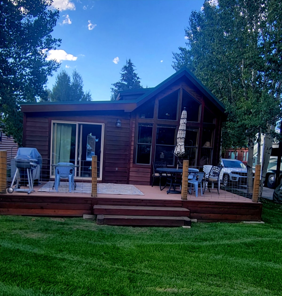 A charming cabin exterior is showcased, featuring a large deck with outdoor seating and a grilling area. The front yard is manicured with lush green grass. Large glass doors provide access to the interior, while abundant trees surround the property, enhancing the natural setting.