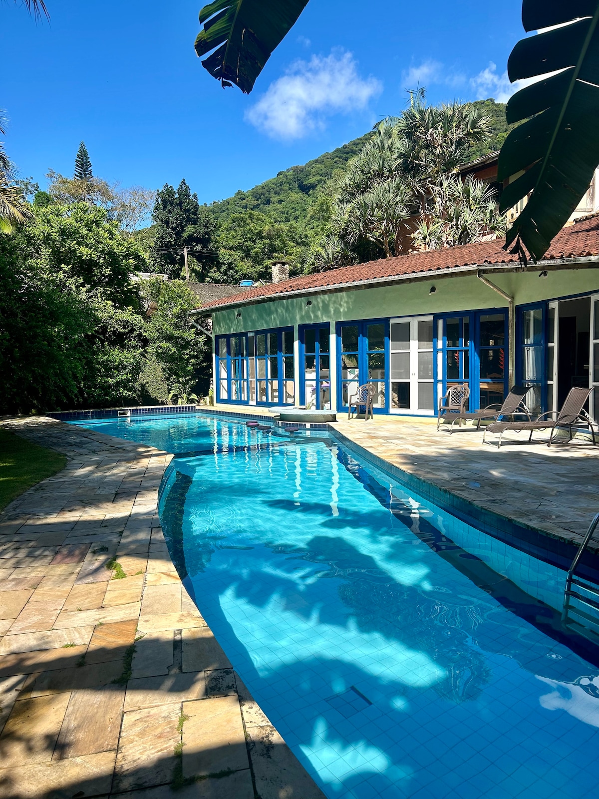 A serene swimming pool is nestled beside a spacious house, with a patio area featuring comfortable lounge chairs. Lush greenery adds to the tranquil setting, while the backdrop of a mountain complements the clear blue sky.