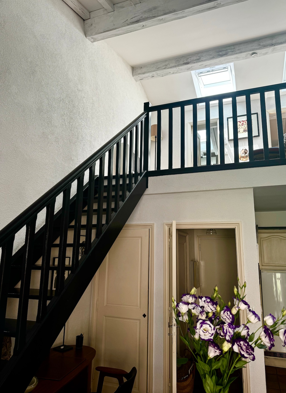 A spacious entryway features a black staircase leading to an upper level. Natural light from a skylight illuminates the white walls, highlighting the textures. A wooden desk is visible, accompanied by fresh flowers in a vase, adding a touch of color to the serene atmosphere.