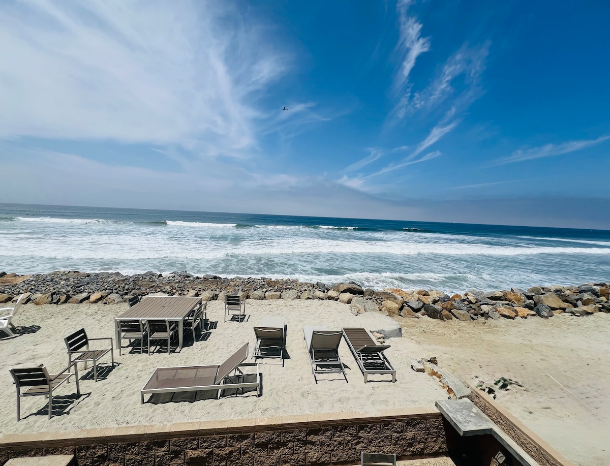 A sandy outdoor area is presented, featuring several lounge chairs and dining tables arranged for outdoor gatherings. The view extends to the ocean, where waves gently crash against the rocky shore under a clear blue sky with scattered clouds.