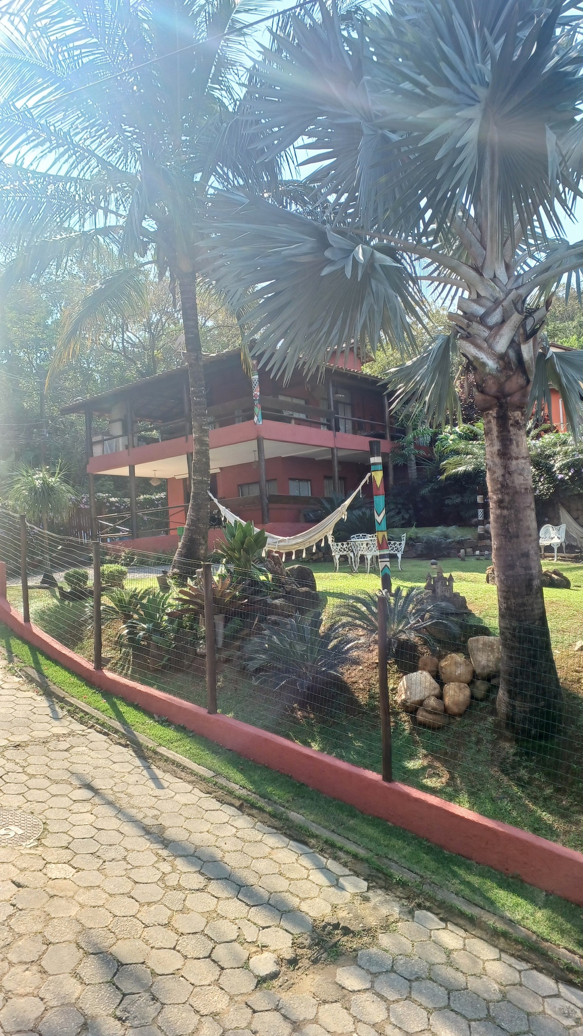 The property is surrounded by lush greenery, featuring palm trees and tropical plants. A two-story building is visible, showcasing a welcoming facade. A pathway leads to the entrance, framed by well-maintained landscaping and decorative elements, creating an inviting outdoor atmosphere.
