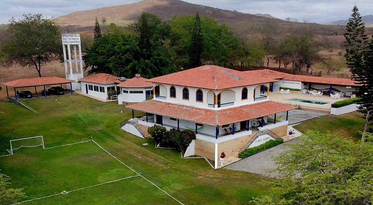The image captures a spacious property featuring a two-story house with a red-tiled roof, surrounded by expansive greenery. Various outdoor spaces, including a soccer field and a terrace area, can be seen. A mix of mature trees provides shade throughout the landscape.