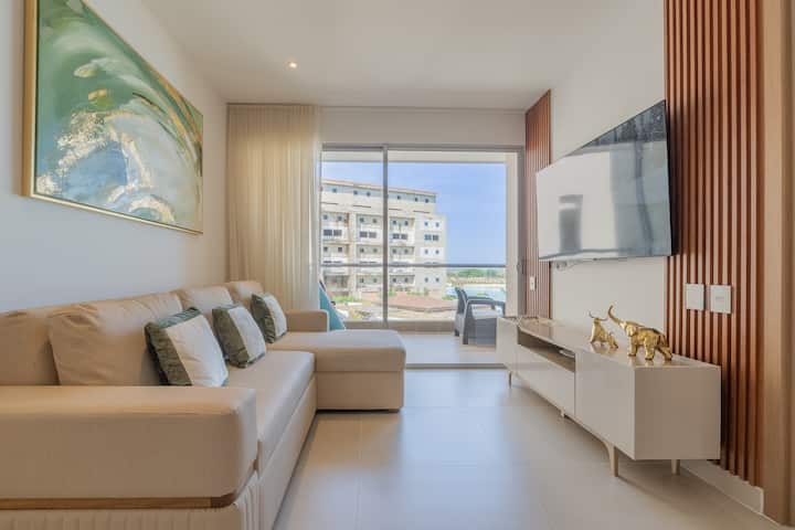 Zona de estar equipada con TV de pantalla grande sobre elegante panel de madera, amplio sofa modular, decoración moderna y acceso directo al balcón con vista exterior.