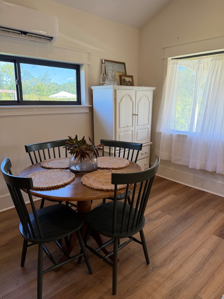 The cottage dining nook with a round wooden table, dark chairs and soft window curtains