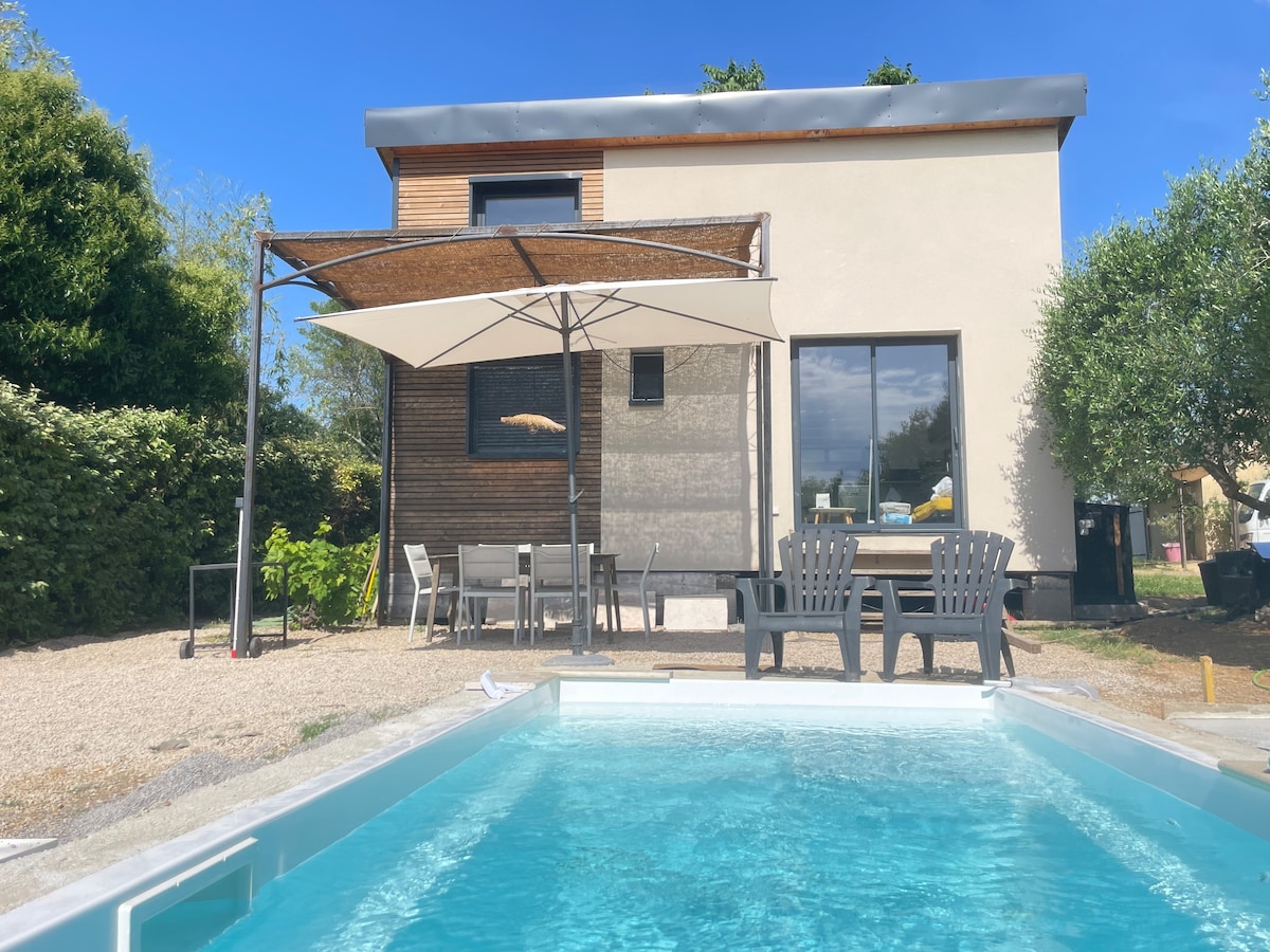 A modern wooden house is displayed, featuring large windows and a shaded seating area with a table and chairs. In the foreground, a private heated pool is visible, surrounded by a gravel area and greenery, creating a serene outdoor environment.