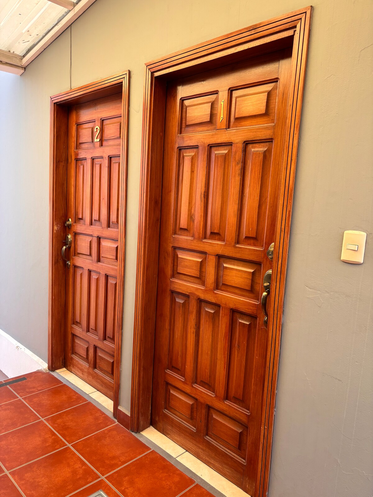 Two wooden doors are positioned along a neutral wall, featuring detailed paneling and gold-colored handles. The floor is tiled in warm terracotta. The door on the left is numbered 2, while the door on the right displays the number 1.