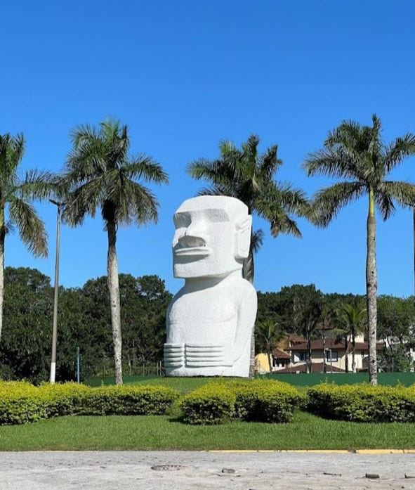 A large white sculpture is prominently displayed in a landscaped area, surrounded by palm trees. Lush green bushes create a vibrant contrast against the blue sky, enhancing the serene outdoor environment. The sculpture serves as a focal point within the picturesque setting.