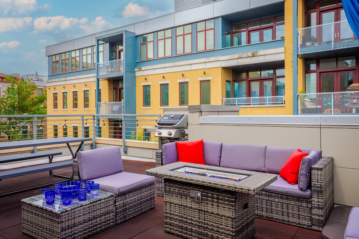 The rooftop patio features a spacious seating area with modern wicker furniture and colorful cushions. A fire table is positioned in the center, surrounded by a glass top, while a natural gas grill is nearby. The backdrop consists of vibrant buildings in the neighborhood.
