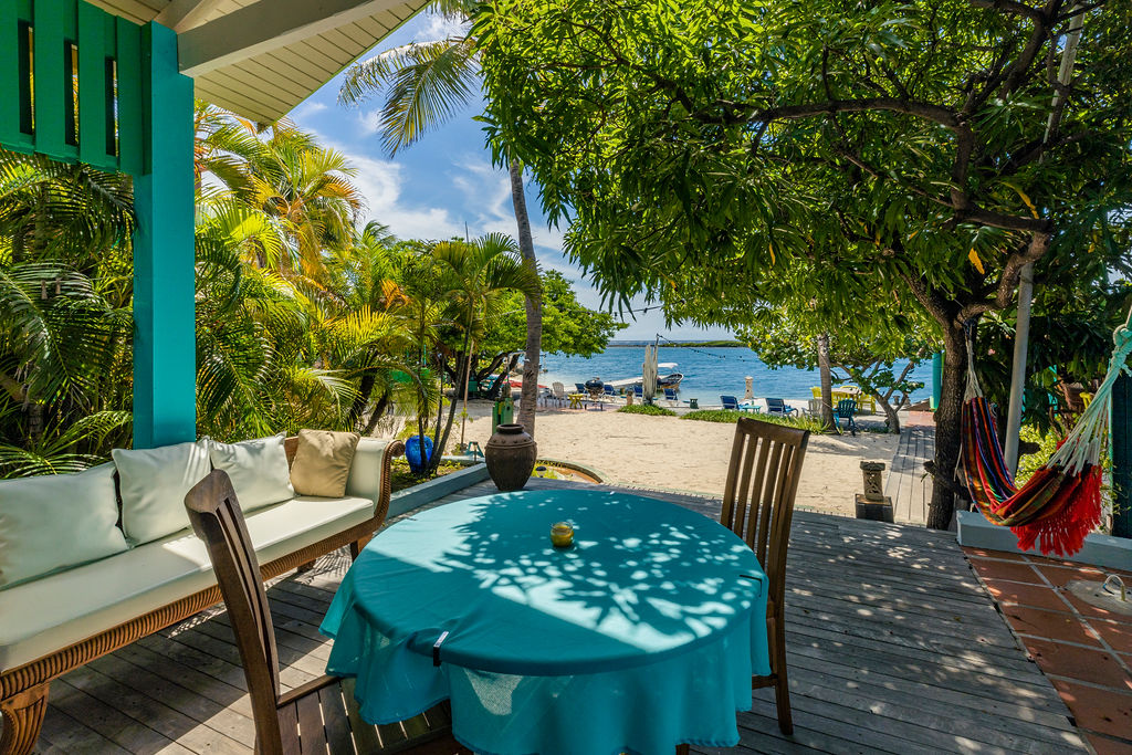 A shaded porch area features a round table draped with a teal tablecloth, surrounded by two wooden chairs. A plush couch is situated nearby, with tropical plants framing the view of the sandy beach and ocean in the distance.