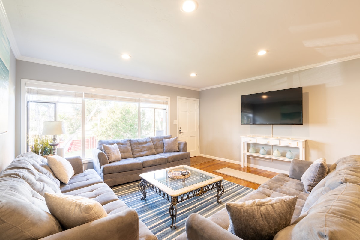 A spacious living room features a large sectional sofa arranged around a coffee table. Natural light floods the area through multiple windows, creating a bright and inviting space. A 50-inch smart TV is mounted on the wall, complementing the modern decor.