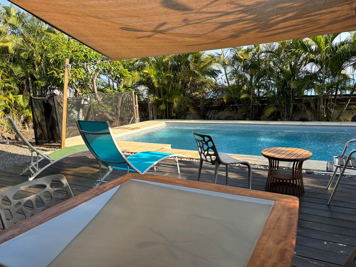 An outdoor terrace features a wooden table and several seating options, including colorful lounge chairs. A spacious pool surrounded by tropical plants is visible in the background, providing a serene setting for relaxation.