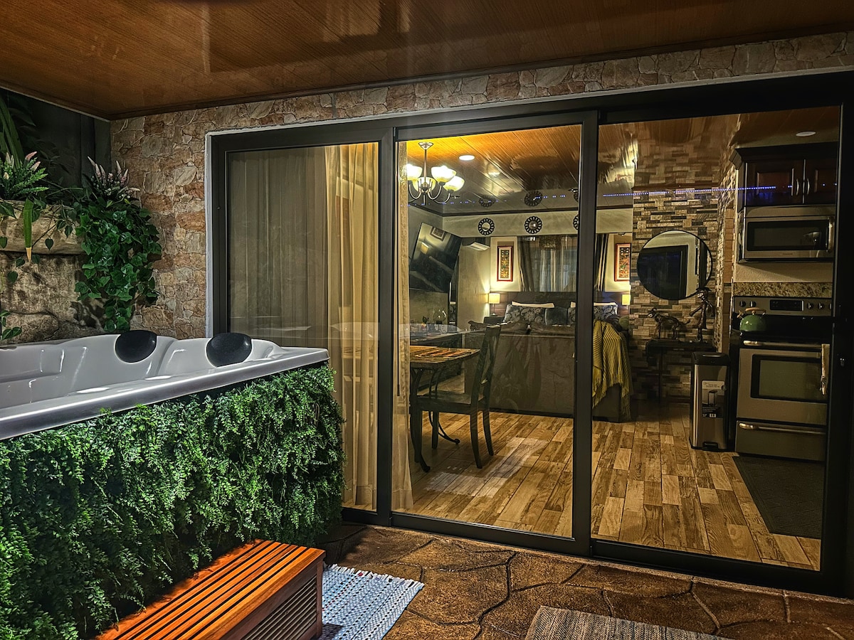 A private hot tub is surrounded by greenery on a patio, providing a sense of tranquility. The glass doors lead to a stylish interior featuring a dining area and a well-equipped kitchen. Warm lighting and natural wood accents create a welcoming ambiance.