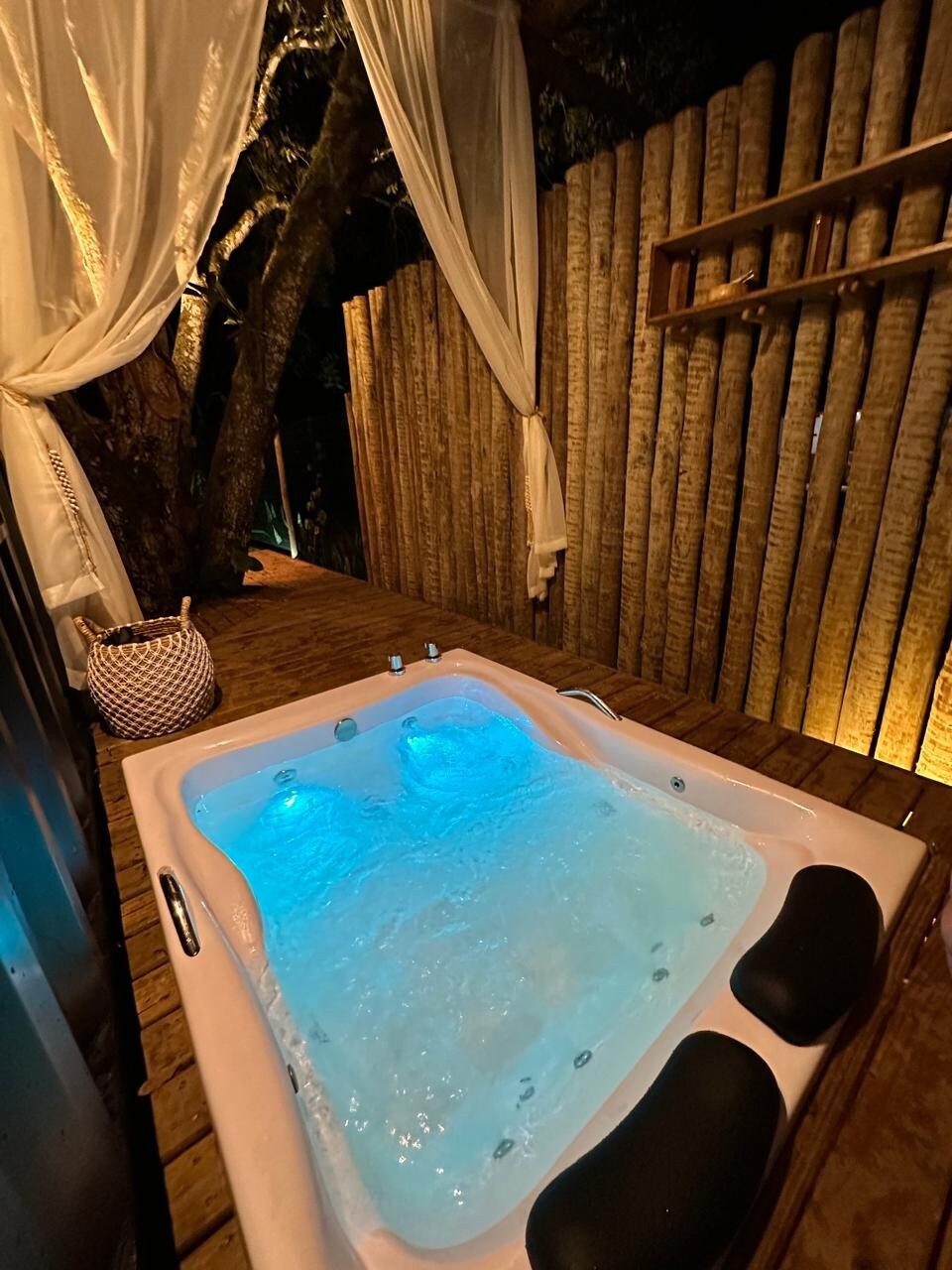 A relaxing hot tub is positioned on a wooden deck, surrounded by softly illuminated wooden fences. Gentle blue lighting enhances the serene ambiance, while sheer curtains provide privacy. The spacious tub features a textured surface for comfort.