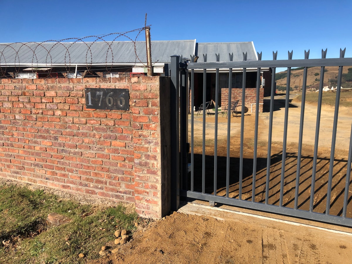 A solid brick wall includes a clear number plaque indicating '1766,' while a metal gate is partially open, leading into the property. The surrounding landscape features open dirt space, with a backdrop of mountains under a clear blue sky.