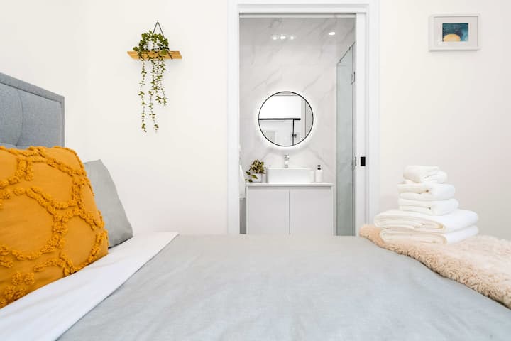 Recorre esta perspectiva de la recámara, mirando desde la ropa de cama gris y mostaza hacia una puerta abierta que conduce a un elegante baño privado con un espejo de pared redondo iluminado.