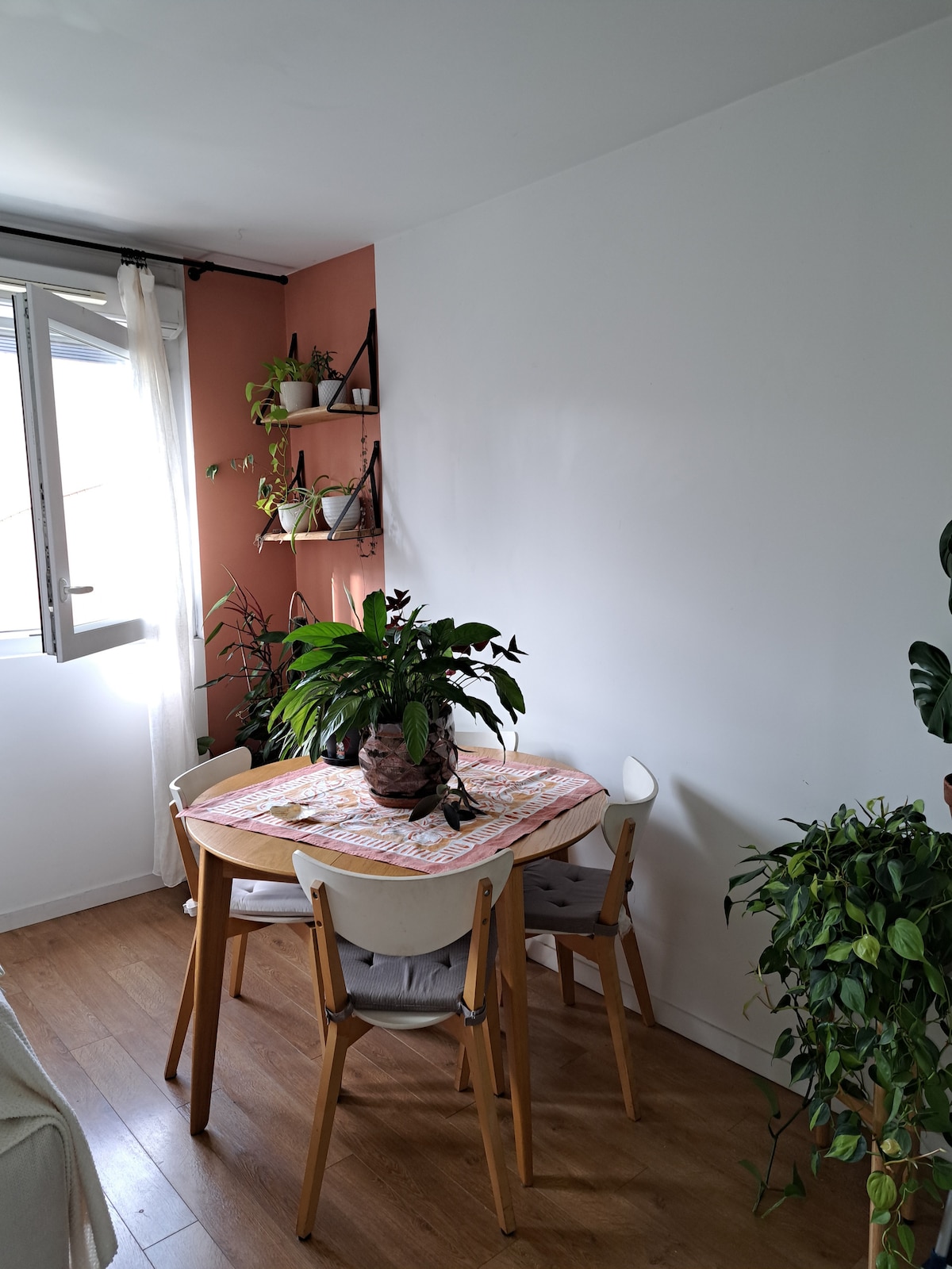 A dining area is showcased with a round wooden table surrounded by four chairs. Natural light enters through a window, illuminating the table adorned with a tablecloth and a plant centerpiece. Greenery is present on shelves and nearby plants, adding a fresh touch to the space.