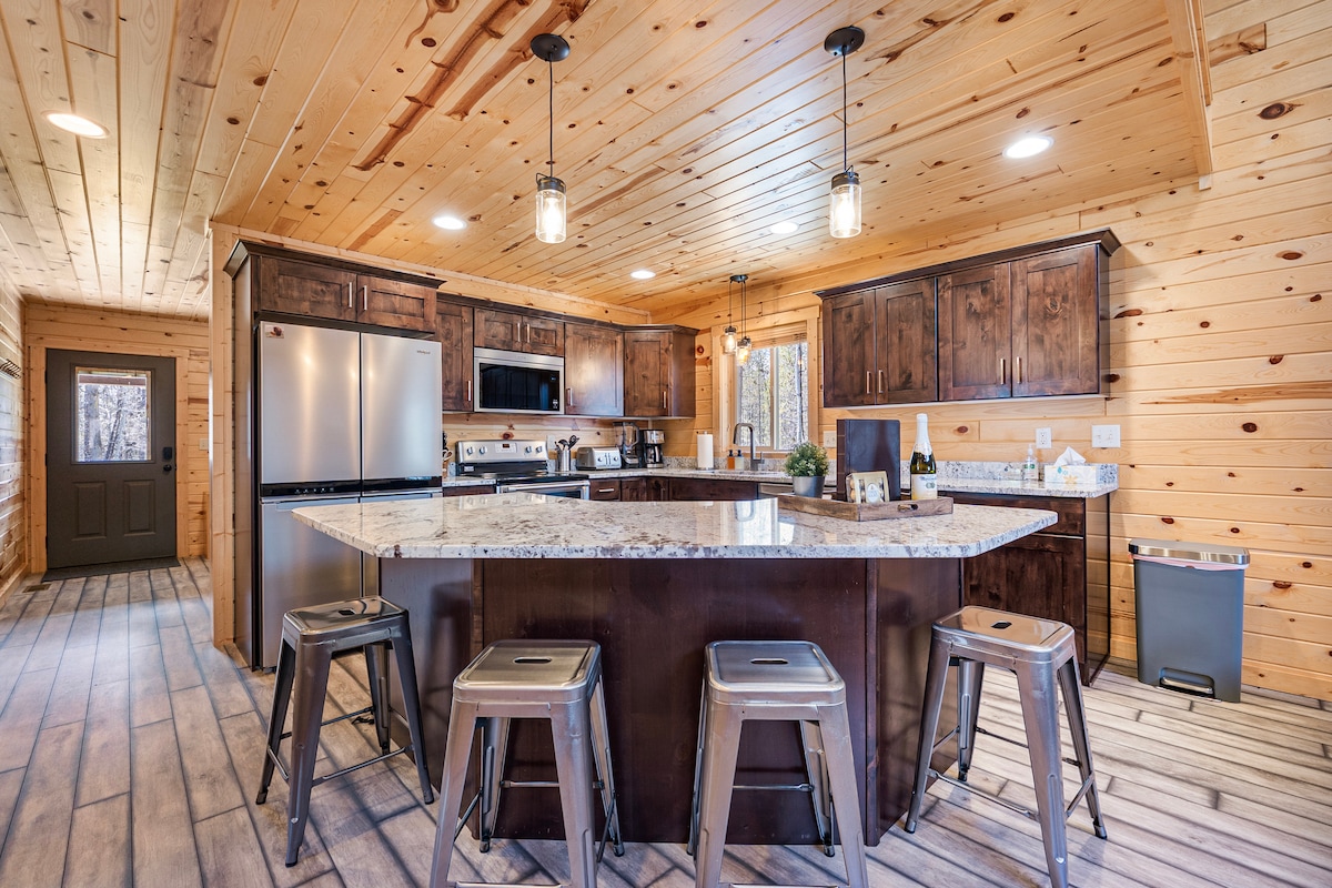 A spacious kitchen features wooden cabinetry and granite countertops, accented by pendant lighting. Stainless steel appliances include a refrigerator and oven. A large island with seating for four is positioned at the center, enhancing the open design. Natural wood walls provide a warm, inviting environment.