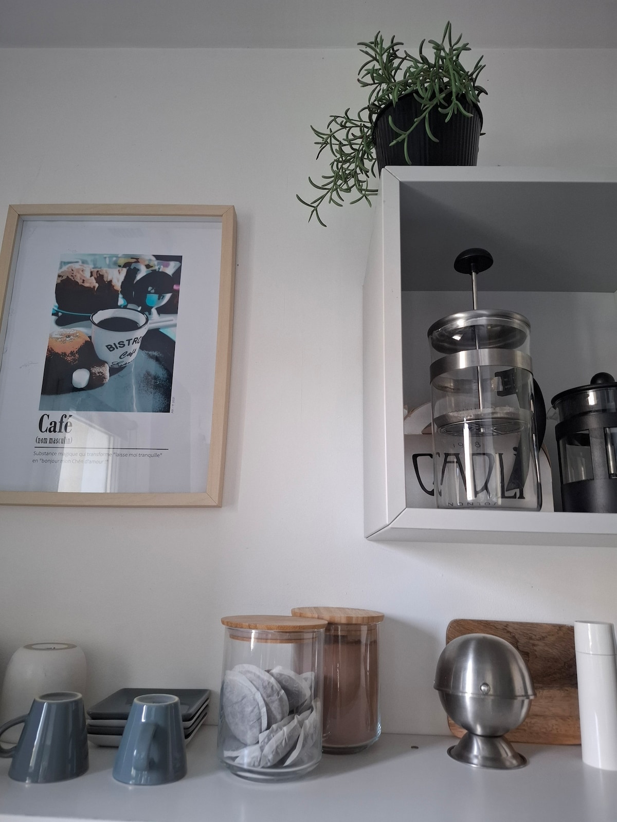 A wall shelf showcases a framed artwork featuring a café scene, accompanied by a small potted plant. Below, clear jars filled with snacks rest alongside wood and metal kitchen accessories, creating an organized and functional space.