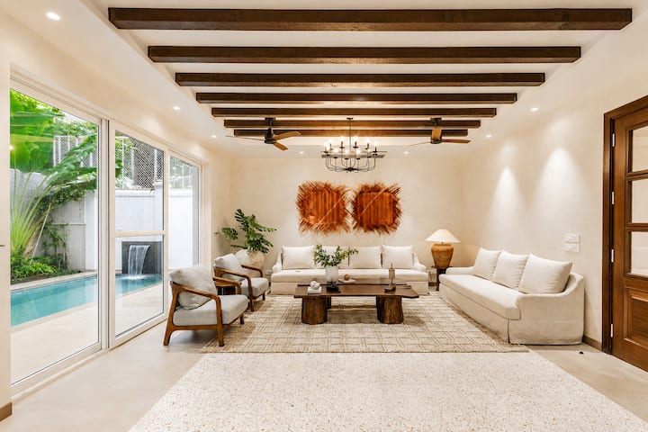 The same room from the front door. Sliding glass runs the length of the pool, so the living area and deck read as one space. The waterfall is visible from every seat.