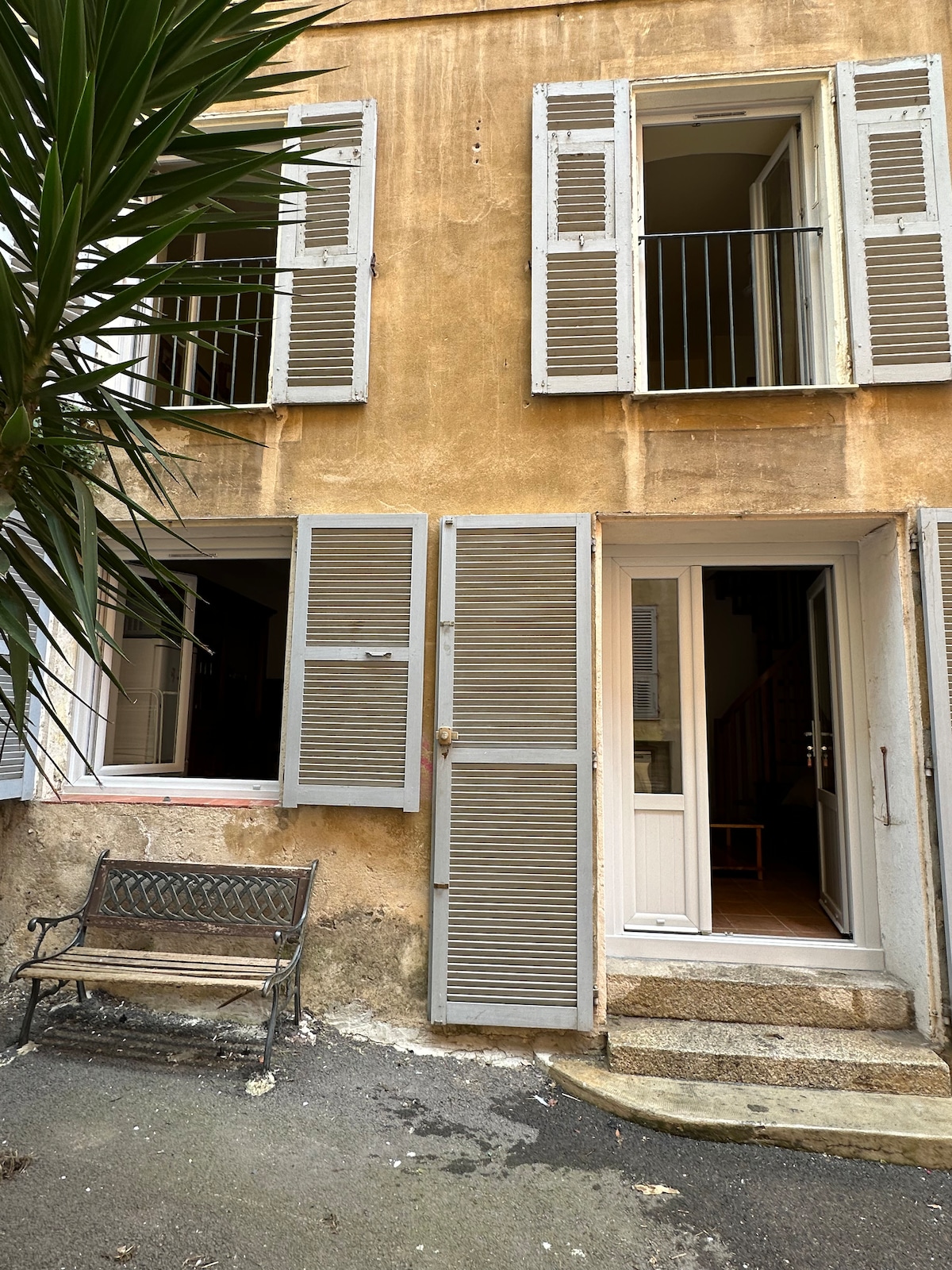 The exterior features a charming façade with light beige walls and traditional shutters, some open to reveal the interior. A small bench is positioned in front of the entrance, framed by a lush palm plant, creating a welcoming entryway.