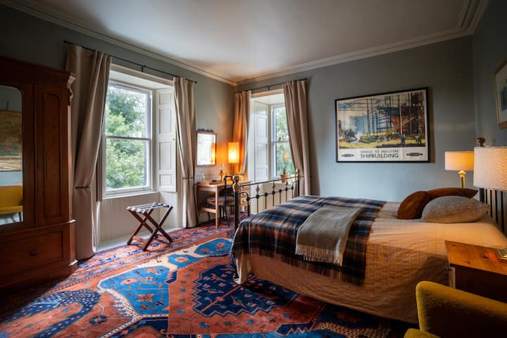 A spacious and cozy bedroom with a queen sized iron bed, two beautiful windows facing the gardens, a clothing closed, sofa and a desk.