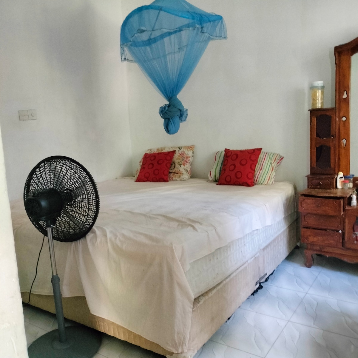 A spacious sleeping area features a large bed with an orthopedic mattress, dressed in neutral bedding and accented by decorative pillows. A ceiling fan is positioned nearby, and a vintage wooden cabinet occupies one corner, providing additional storage.