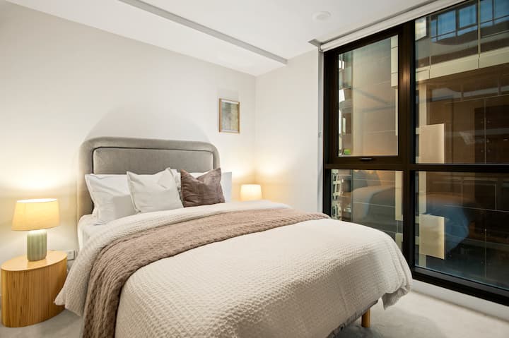 Chambre lumineuse avec un grand lit, une tête de lit rembourrée gris pâle, de la literie blanche et un plaid aux tons neutres chauds. Les lampes de chevet et les grandes baies vitrées créent un cadre confortable et moderne.