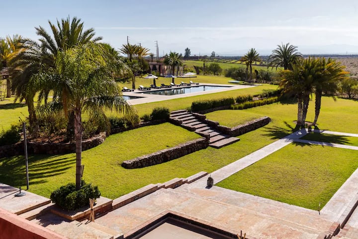 Elevated view across Villa Tameslohte's terraced lawns, palm trees, stone paths and swimming pool towards the Atlas