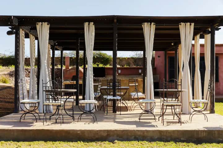 Open-sided pergola at Villa Tameslohte with linen curtains, wrought-iron chairs and the summer kitchen behind
