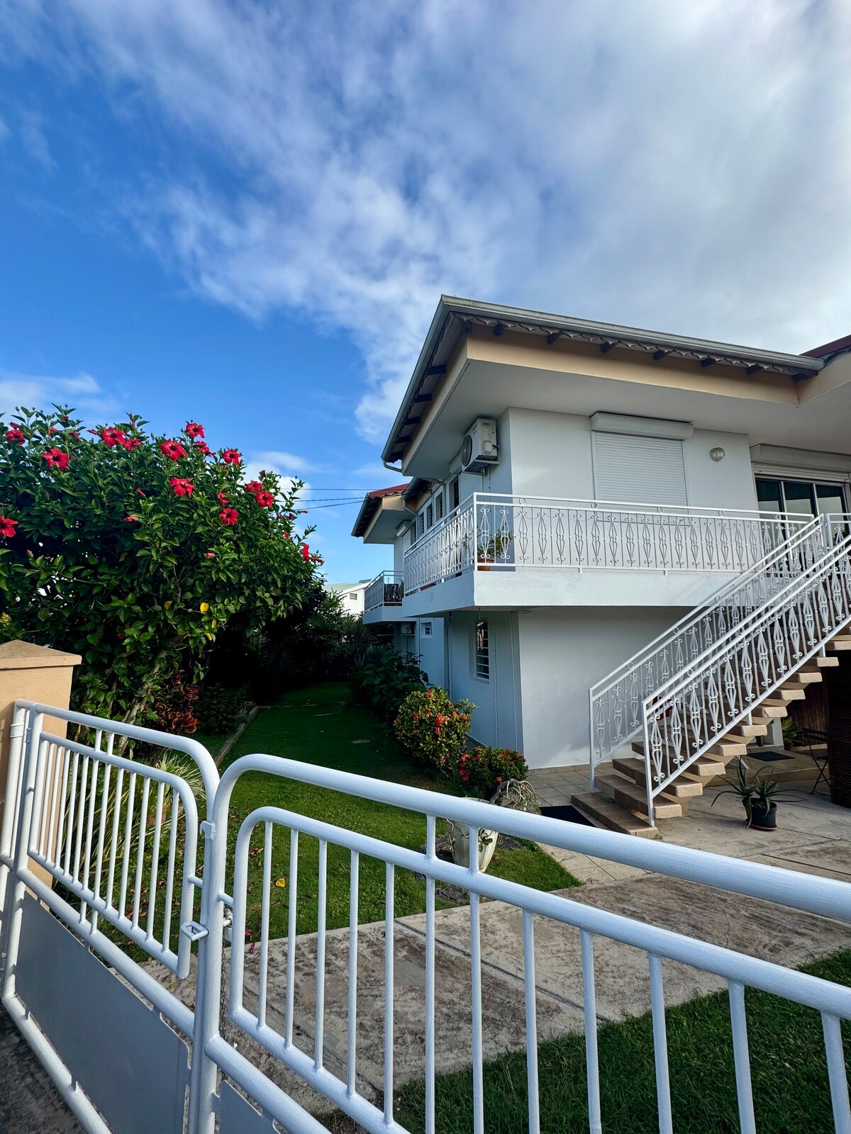 A well-maintained two-story building is shown, featuring a staircase leading to an upper level. Lush greenery, including flowering plants, is visible in the surrounding garden, contributing to a pleasant outdoor space. A clear blue sky adds to the welcoming atmosphere.