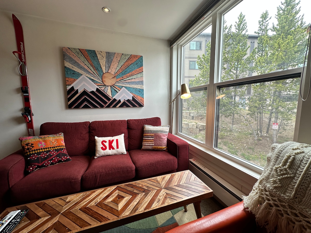 A cozy living area features a red sofa adorned with decorative pillows, including one with the word 'SKI.' Natural light floods the space through large windows, showcasing a view of trees outside. A wooden coffee table with a geometric design sits in front of the sofa.